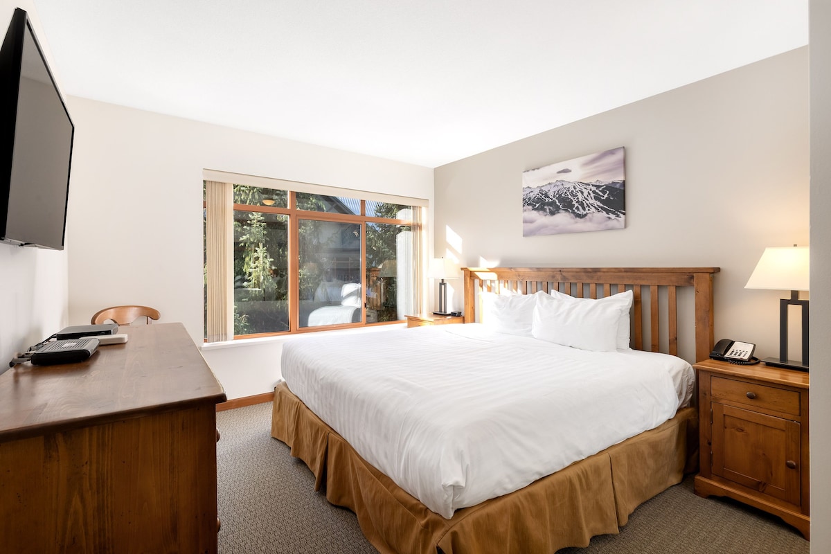 A hotel room with a large bed, a desk, a chair, a TV, a window with a view of trees, and a painting on the wall.