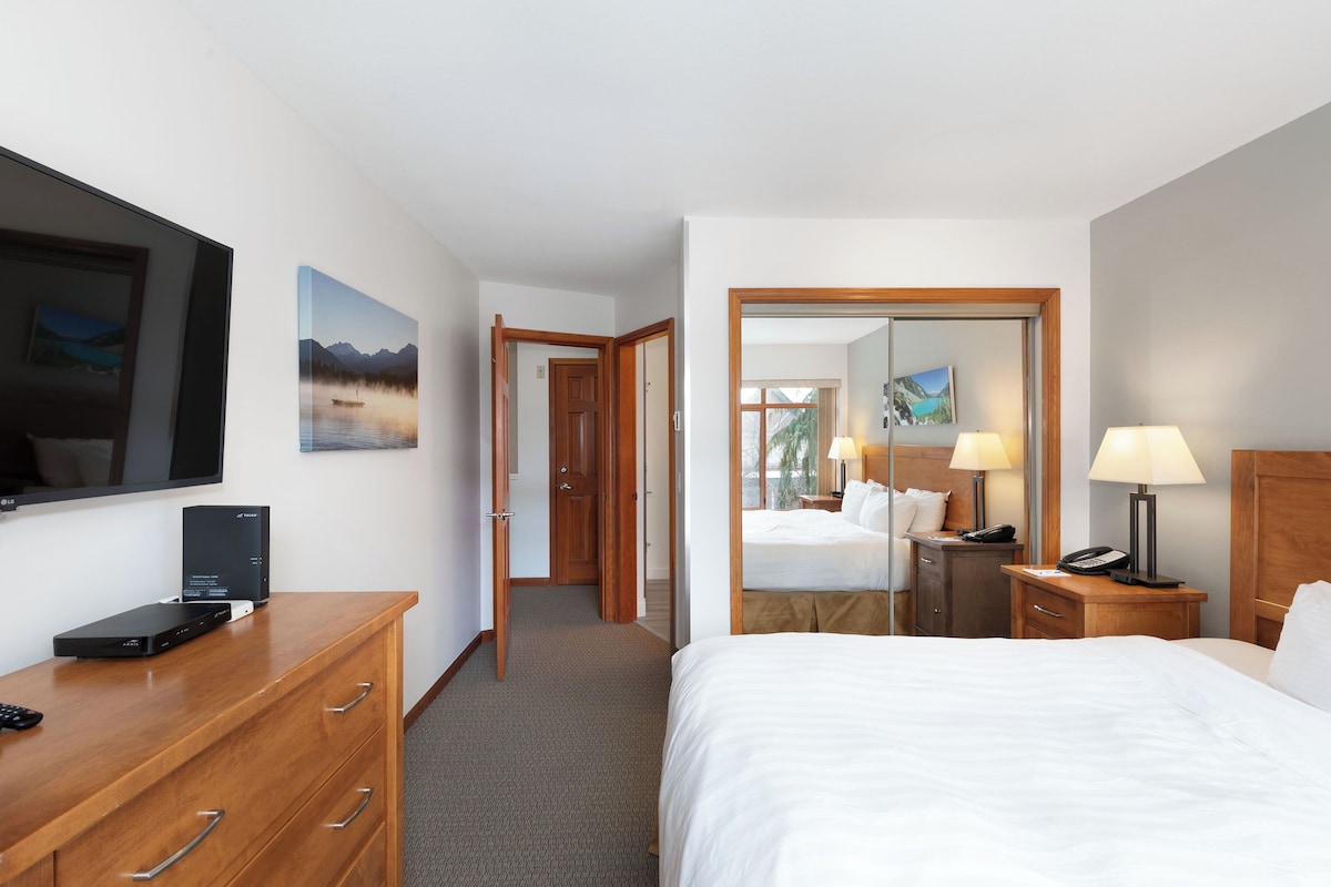 A hotel room with a bed, a desk with a TV, a dresser, a mirror, and a door leading to another room.