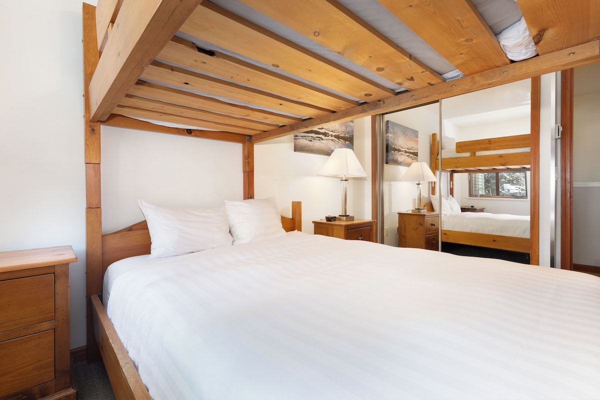 A bedroom with a wooden bed frame, white bedding, two bedside tables with lamps, and a view of the outdoors through a window.