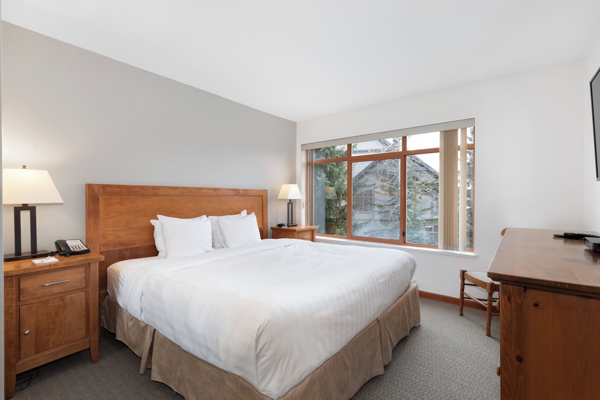 A hotel room with a large bed, bedside tables with lamps, a desk, and a window with a view of trees and buildings.