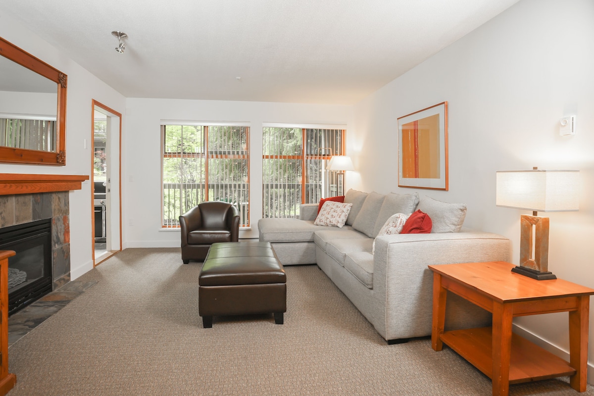 A living room with a fireplace, a sofa, an ottoman, and a table with a lamp.