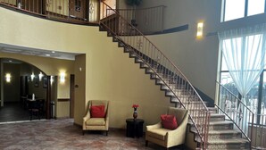 Um hall espaçoso com teto alto, escada com corrimão de ferro forjado e área de estar com duas poltronas e um sofá.