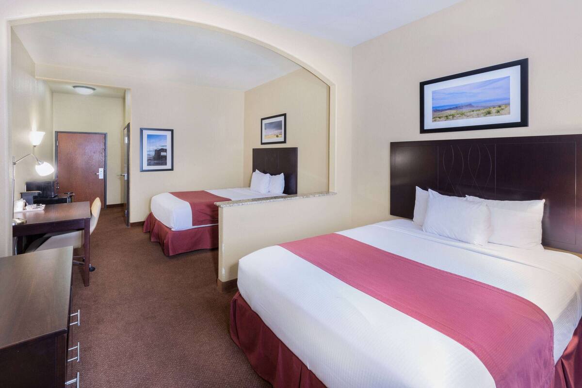 Une chambre d’hôtel avec un grand lit, un lit plus petit, un bureau avec une lampe et une porte donnant sur une autre pièce.