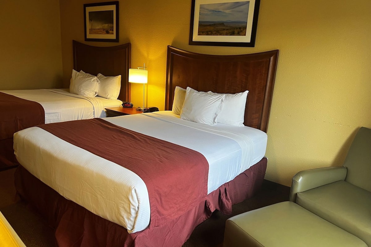 Une chambre d’hôtel avec deux lits, une table de chevet avec une lampe, une chaise et deux tableaux encadrés au mur.