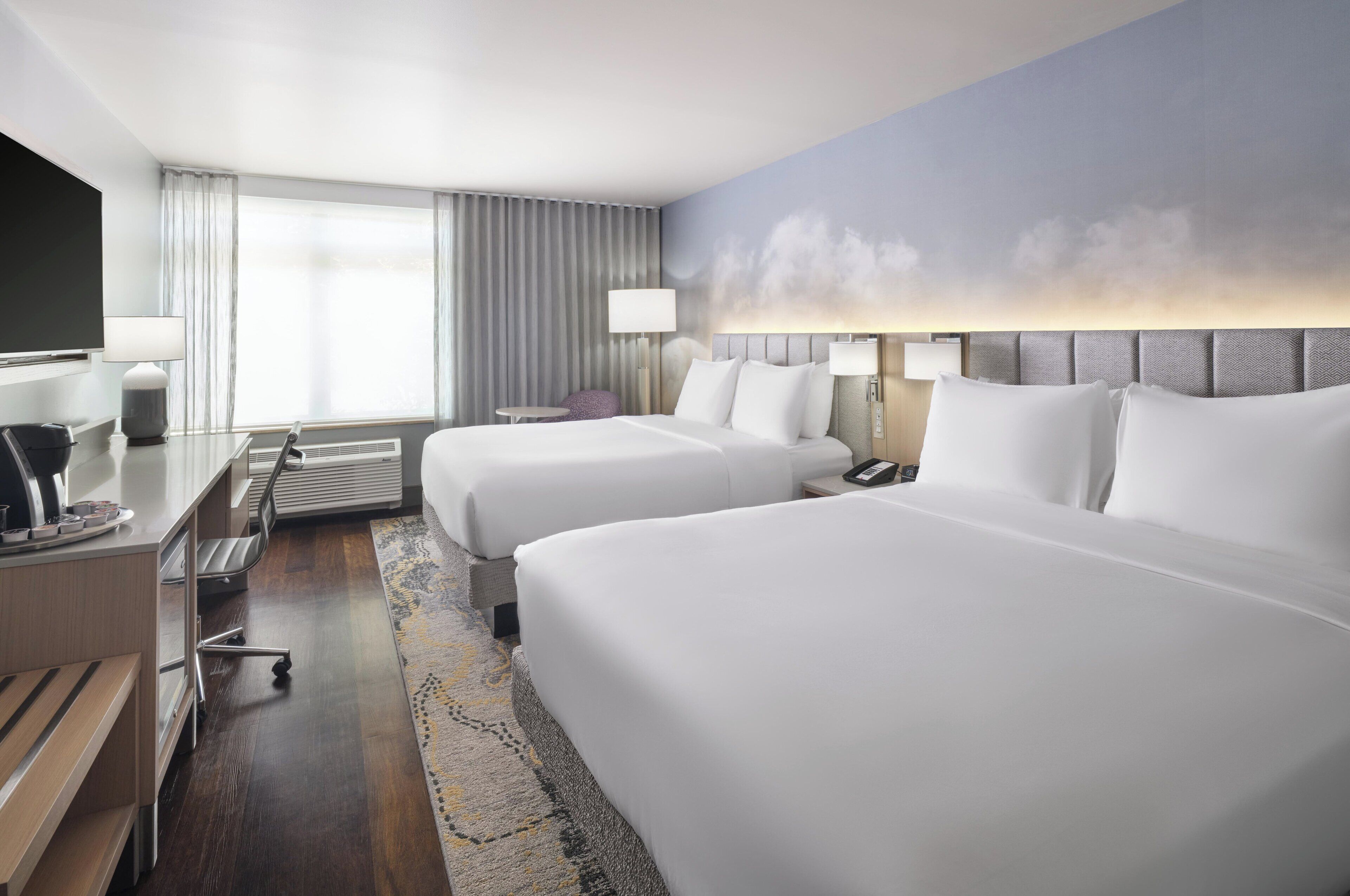 A hotel room with two beds, a desk with a chair, a TV, and a large window with curtains.