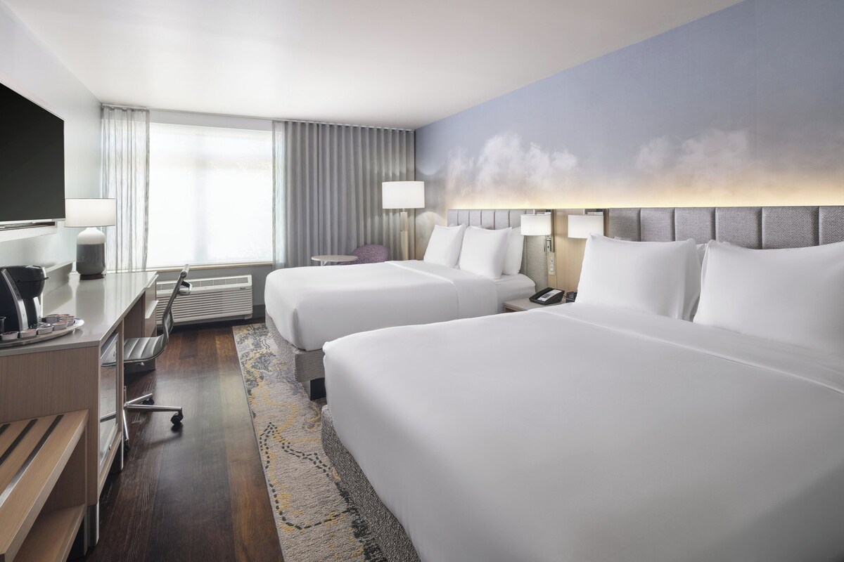 A hotel room with two beds, a desk with a chair, a TV, and a large window with curtains.