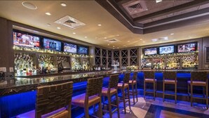 A bar area with multiple screens displaying content, a variety of bottles on shelves, and several bar stools. - Hotel Indigo Long Island - East End by IHG (Riverhead)
