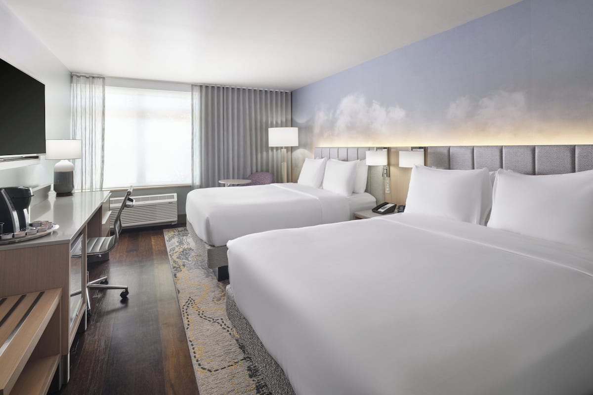 A hotel room with two beds, a desk with a chair, a TV, and a large window with curtains.