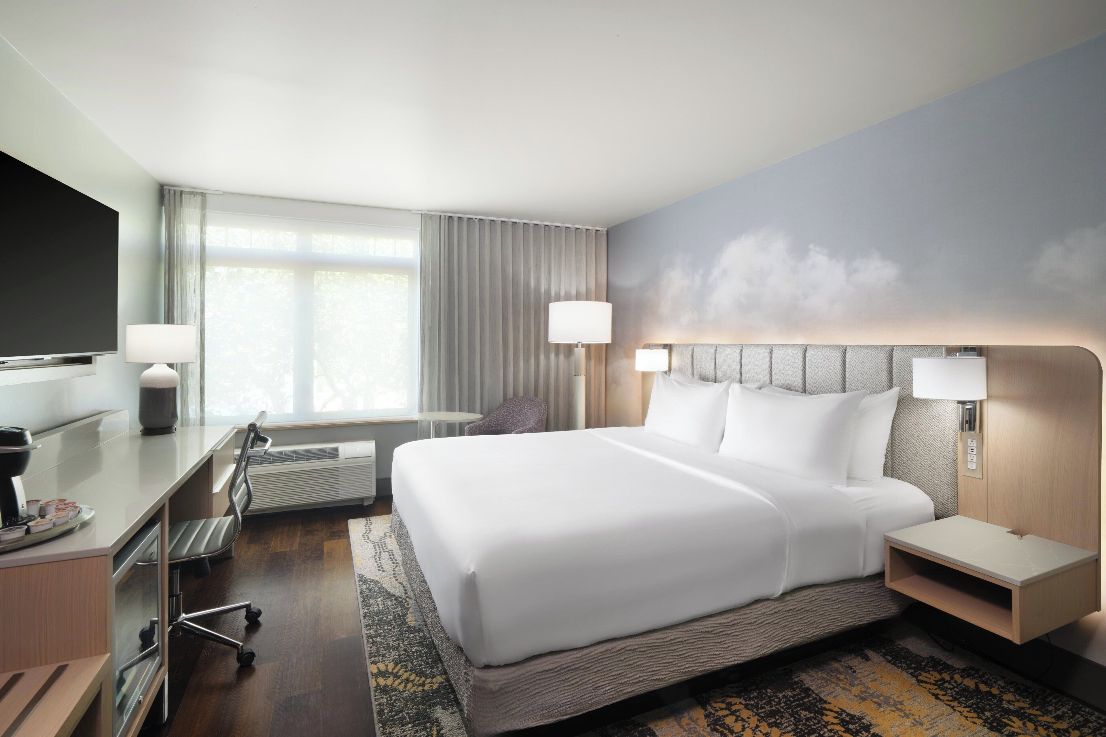 A hotel room with a large bed, bedside tables, a desk with a chair, a TV, and a window with curtains.