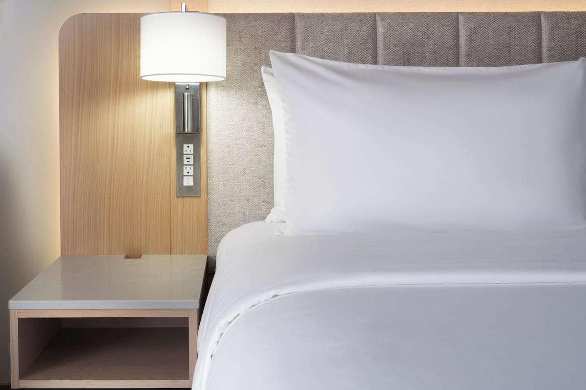 A neatly made bed with white linens, a wooden nightstand with a lamp, and a power outlet.