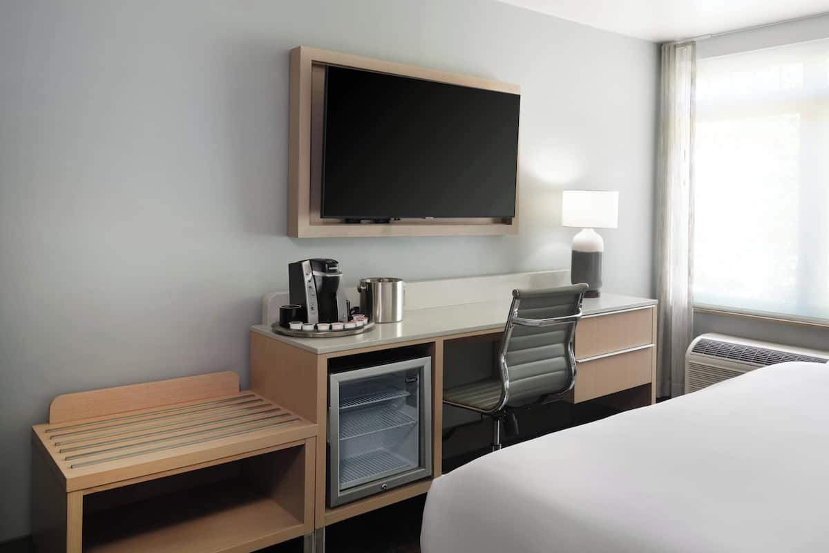 A hotel room with a bed, a desk with a coffee maker, a mini-fridge, a TV mounted on the wall, and a window with curtains.