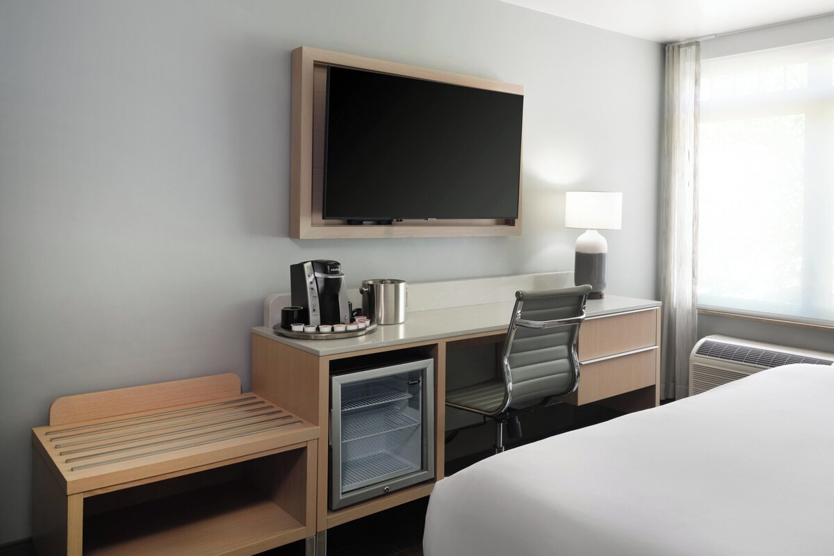 A hotel room with a bed, a desk with a coffee maker, a mini-fridge, a TV mounted on the wall, and a window with curtains.