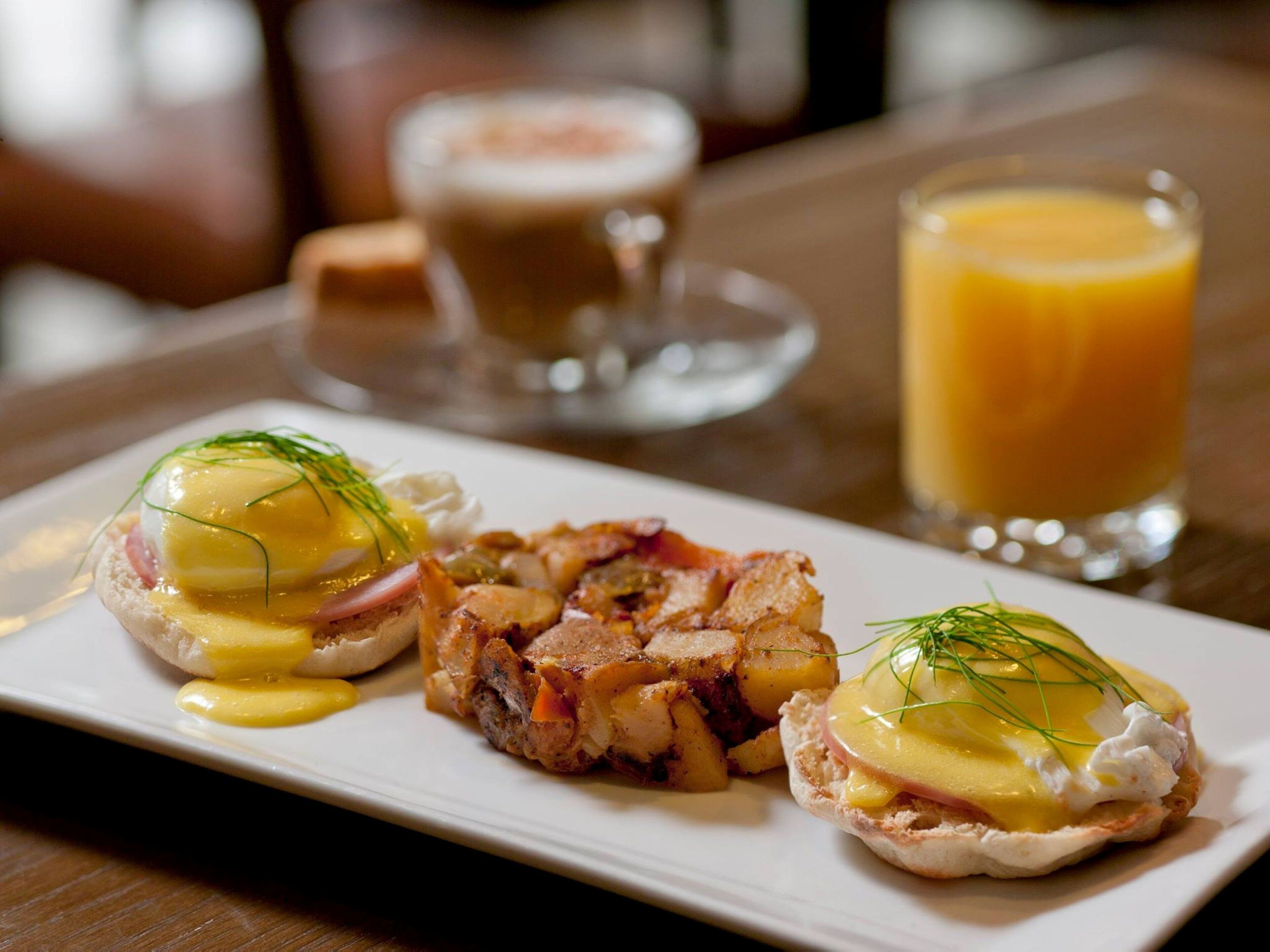Un plato de desayuno con huevos benedictinos y tocino, un vaso de jugo de naranja y una taza de café sobre una mesa de madera.