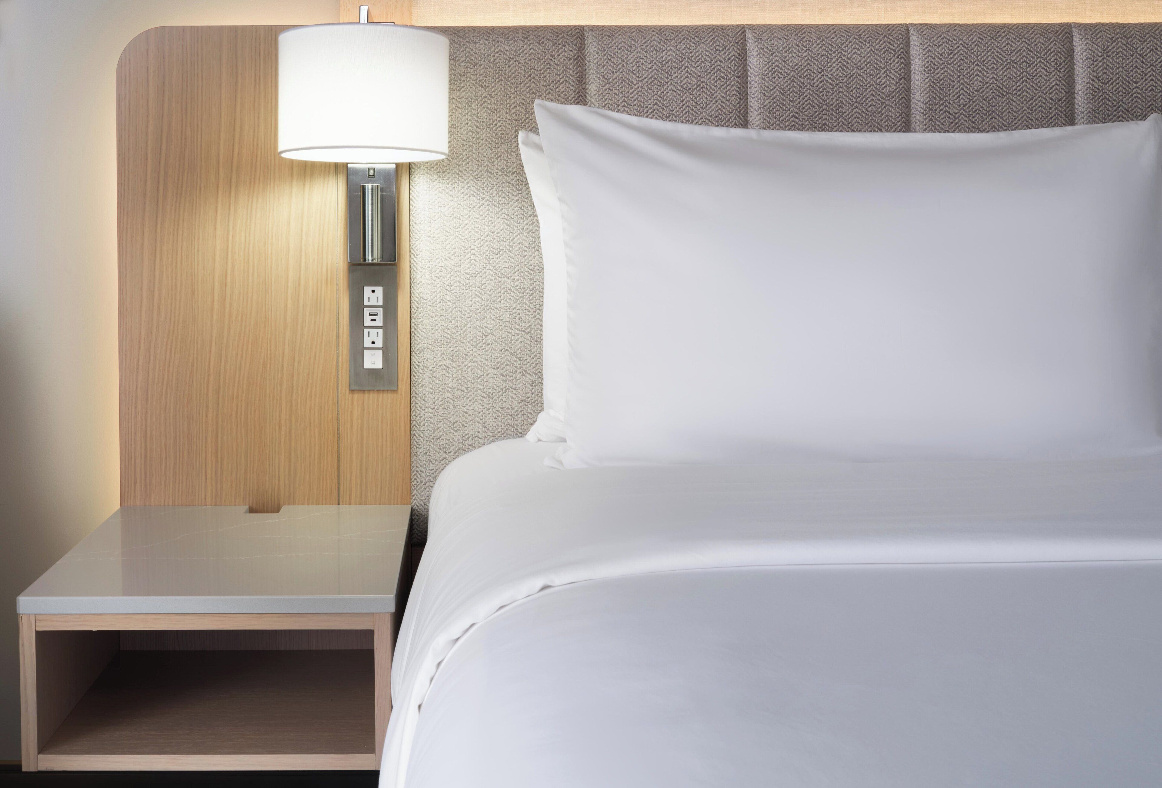 Una cama bien hecha con sábanas blancas, una mesita de noche de madera con una lámpara y un enchufe.