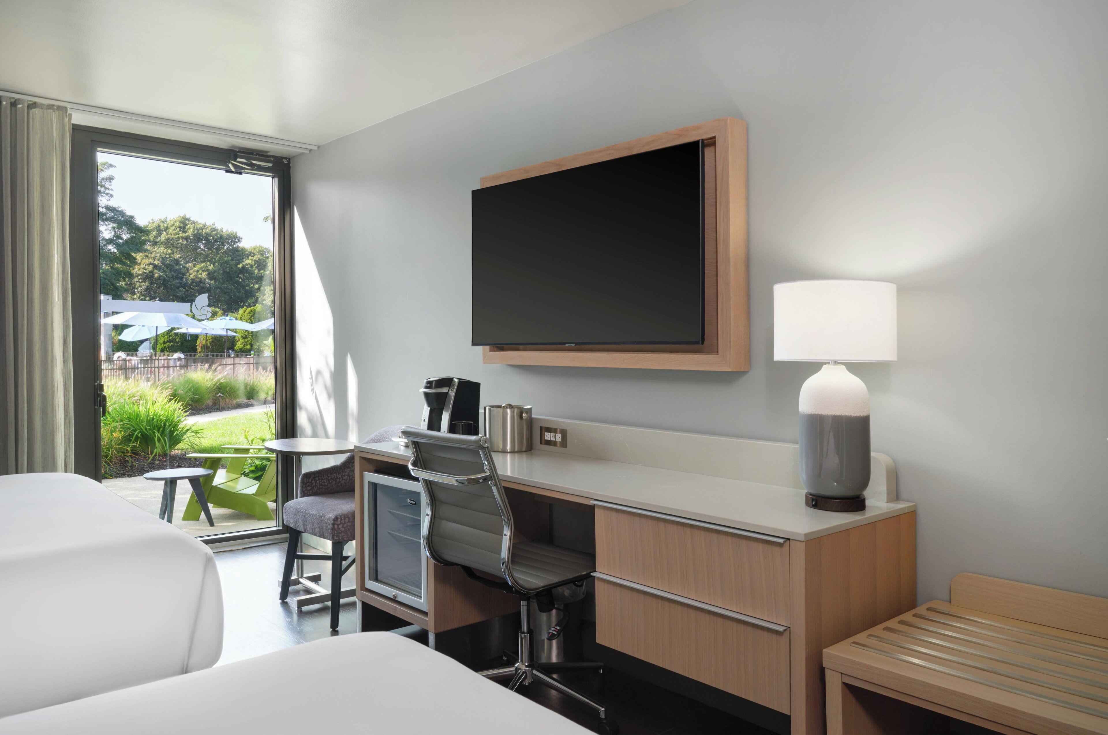 A hotel room with a desk, chair, TV, lamp, and a view of the outdoors.