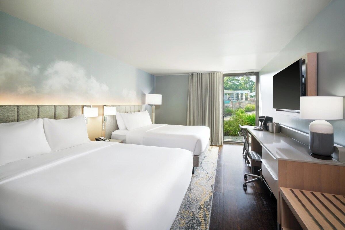 A hotel room with two beds, a desk with a flat-screen TV, a lamp, and a view of greenery outside.