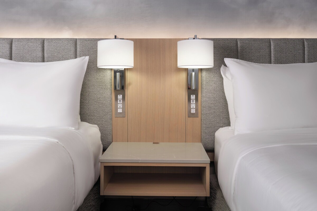 A hotel room with two beds, a wooden headboard, bedside lamps, and a small wooden table.