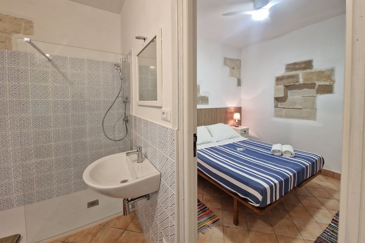 A bathroom with a sink, shower, and patterned tiles. A bed with a striped bedspread is visible in the adjacent room.