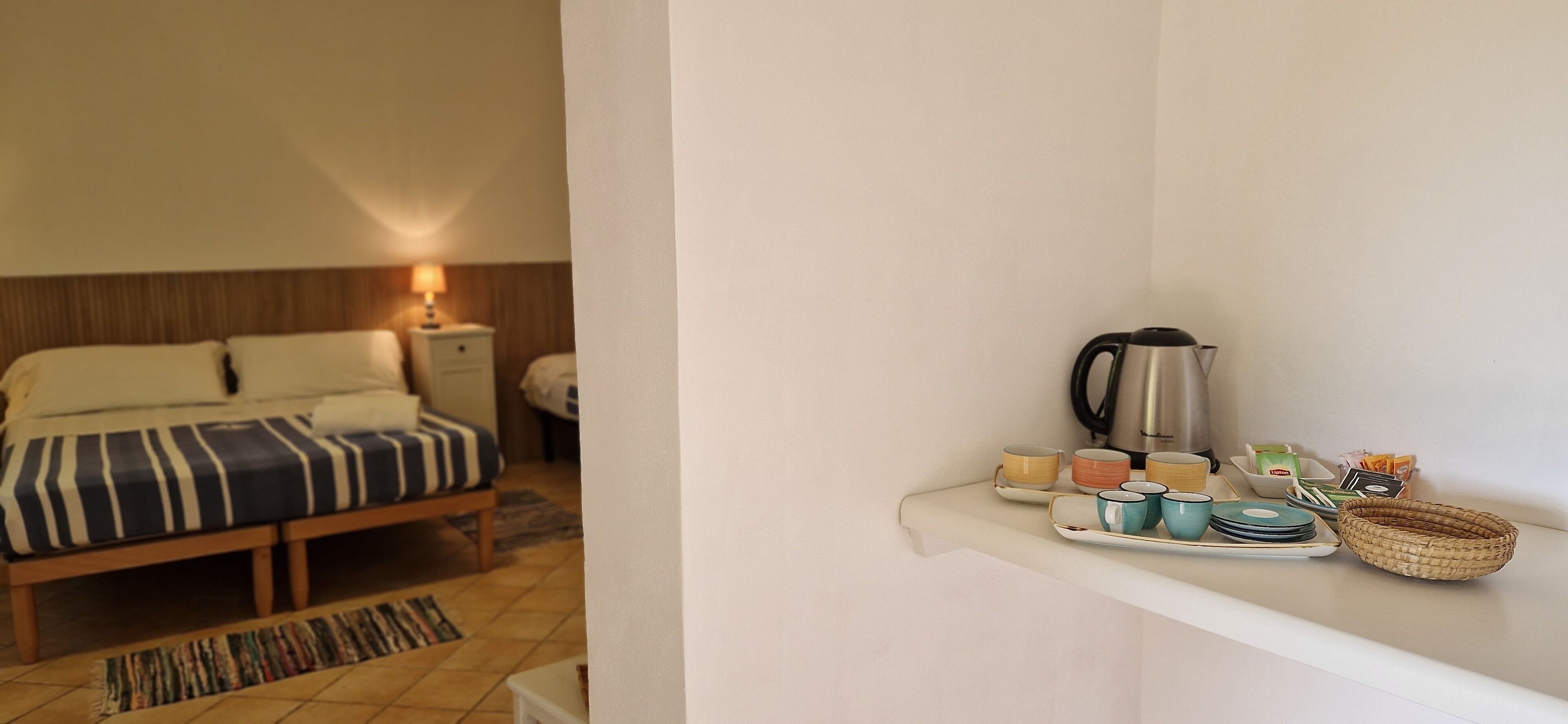 A hotel room with a bed, bedside table, lamp, and a tray with cups and a kettle.