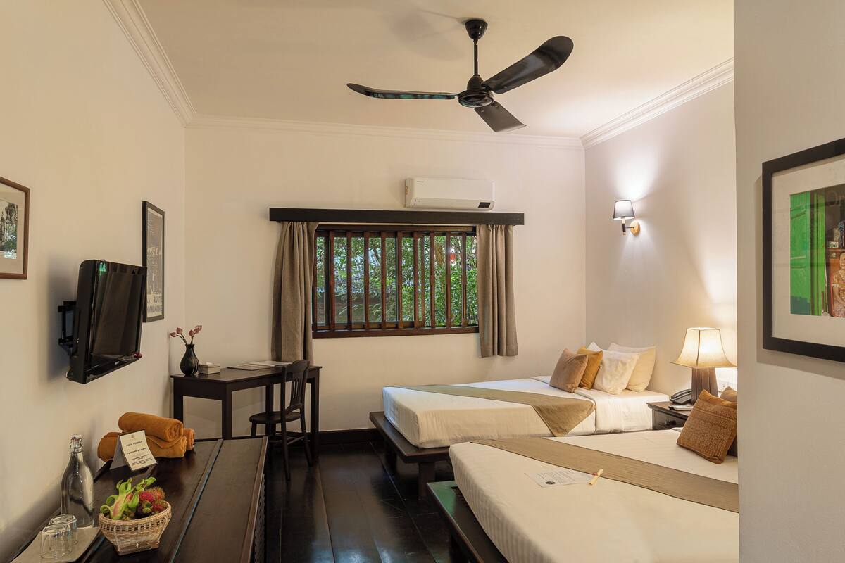 A hotel room with two beds, a ceiling fan, an air conditioning unit, a desk with a chair, a TV, and a window with curtains.