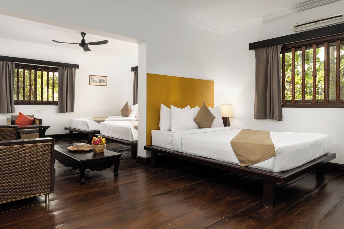A hotel room with a bed, a sofa, a small table with fruit, a wicker chair, and a ceiling fan.