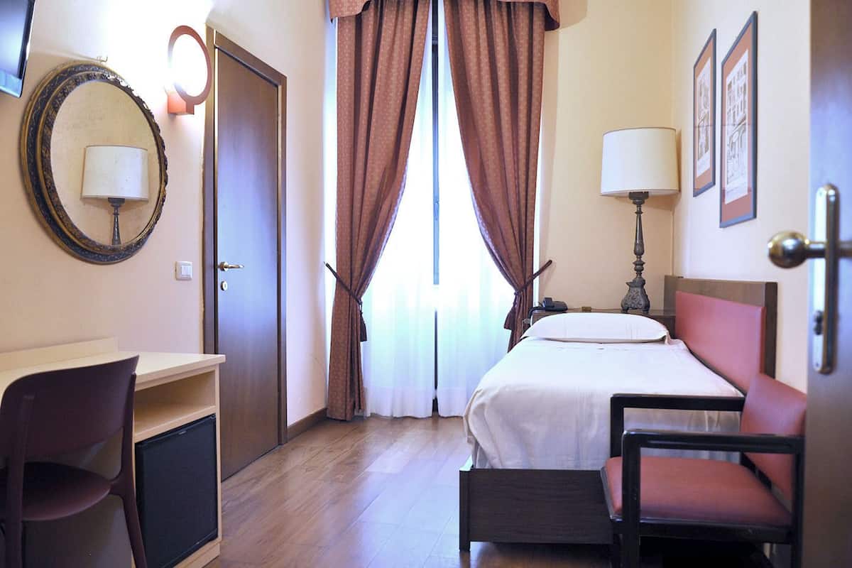 A hotel room with a bed, a desk, a chair, a lamp, and a door.
