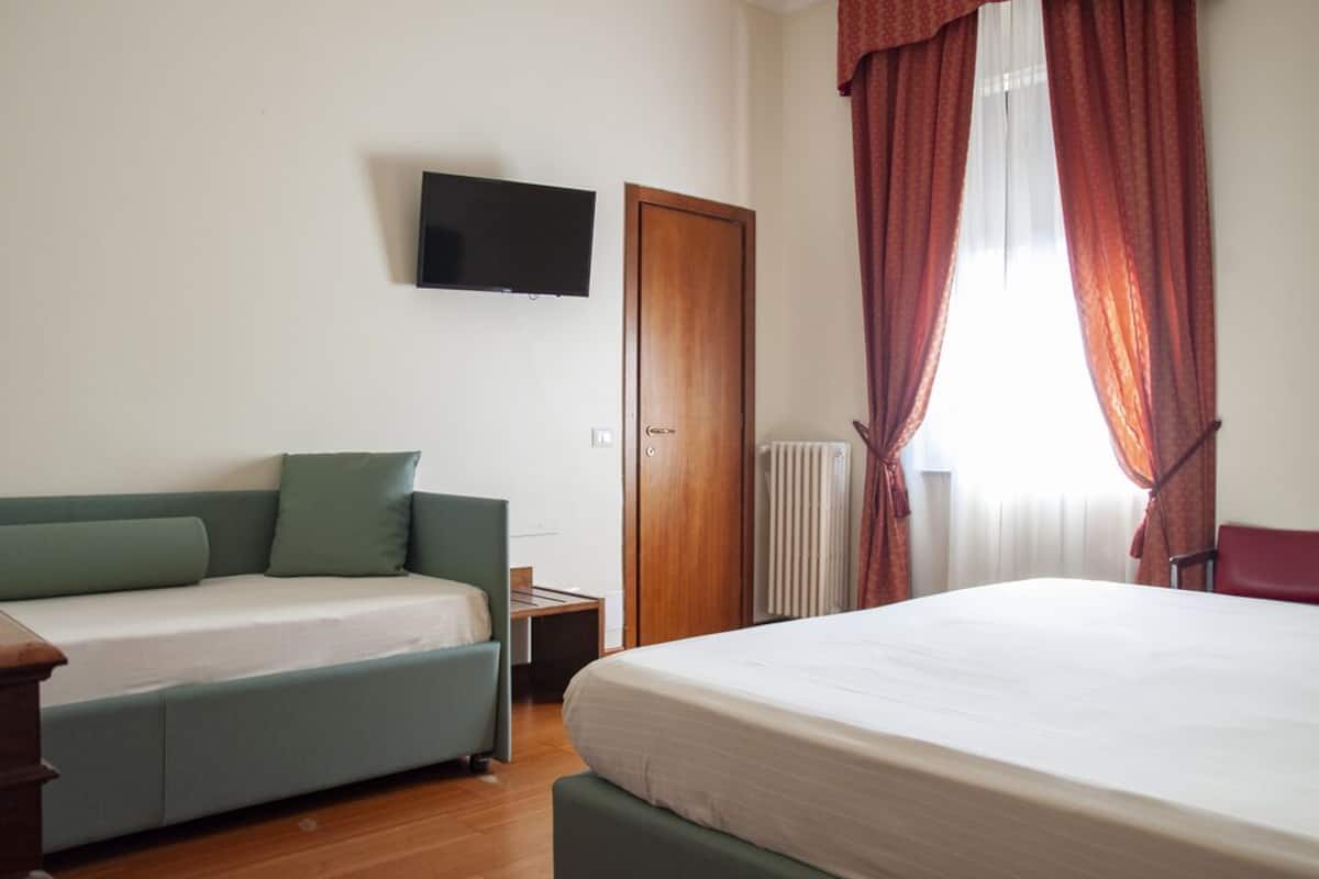 A room with a bed, a sofa, a TV mounted on the wall, a wooden door, and a window with curtains.