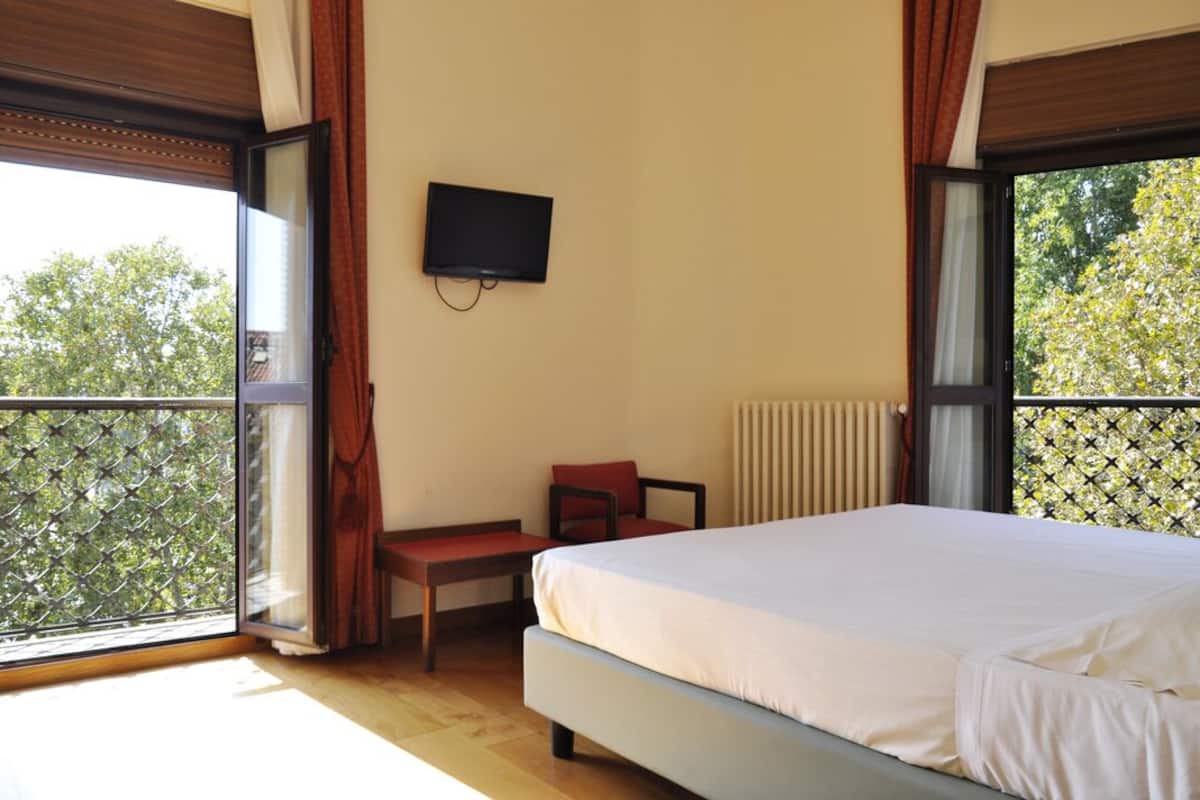 A bedroom with a bed, a TV mounted on the wall, a chair, and a window with a view of trees.