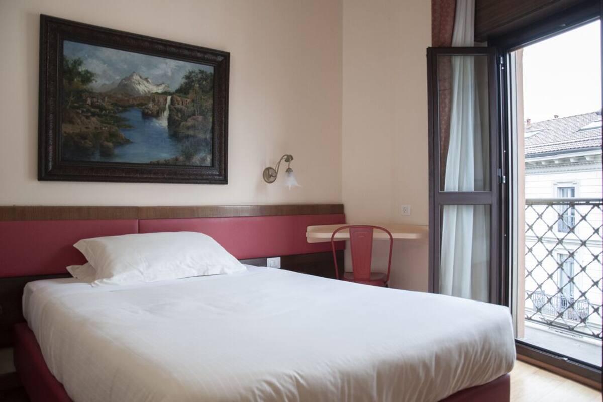 A hotel room with a bed, a painting on the wall, a window with a view of buildings, and a chair.