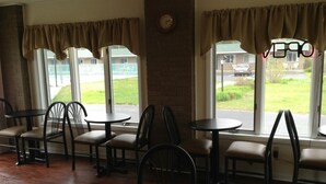 A restaurant interior with multiple tables and chairs, large windows with curtains, and a brick wall. - Capri Motel (Mackinaw City)