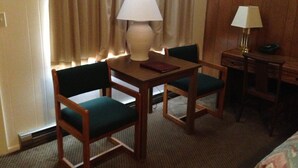 A hotel room with a wooden desk, two green upholstered chairs, a white lamp on the desk, and a lamp on the side table. - Capri Motel (Mackinaw City)