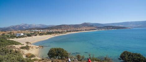 A coastal landscape with a beach, mountains, and two cyclists on a trail.