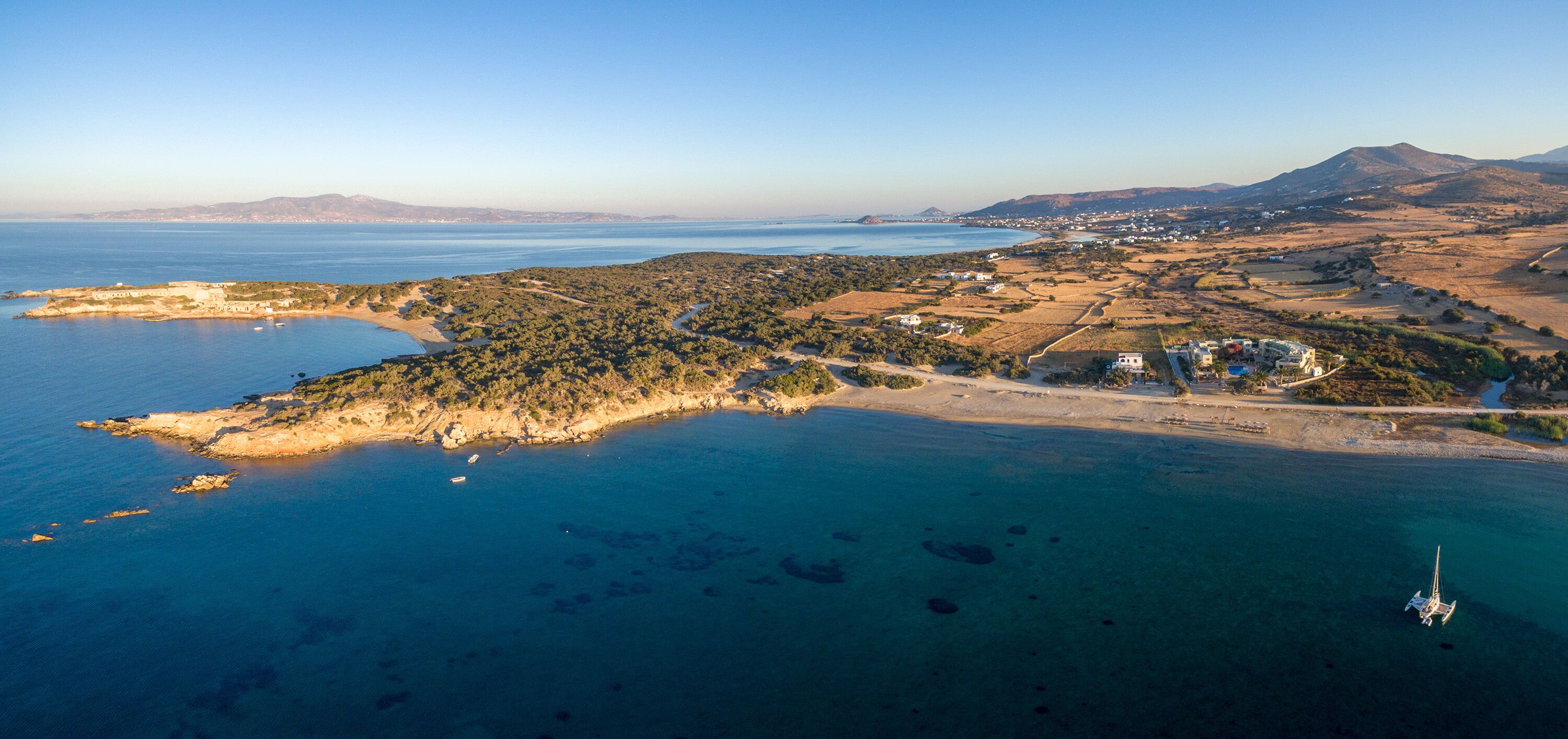An aerial view of a coastal area with a small island, clear blue waters, and a distant mountain range.