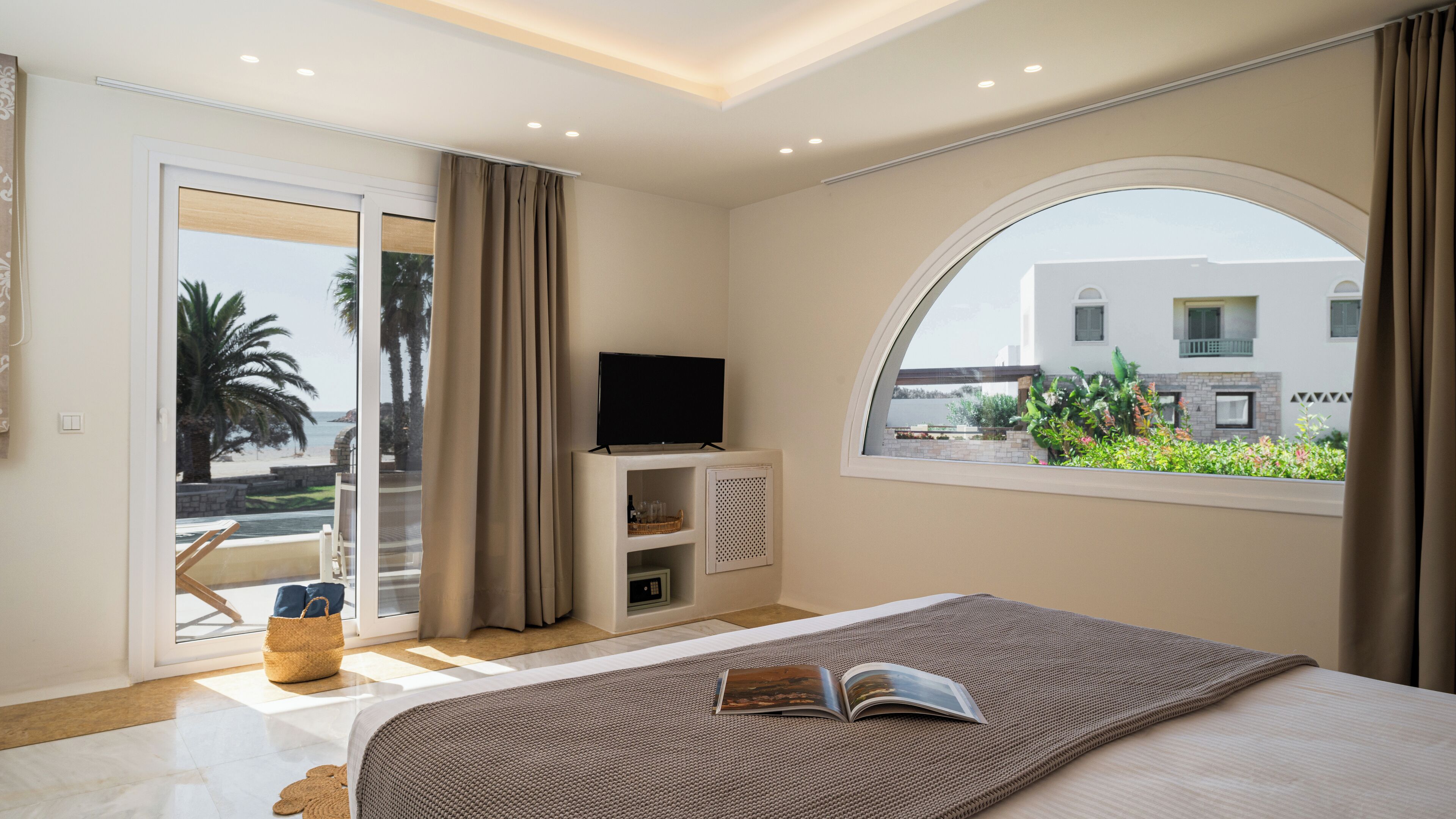 A bedroom with a large bed, a TV on a stand, a window with a view of a building and greenery, and a balcony with a chair and a basket.