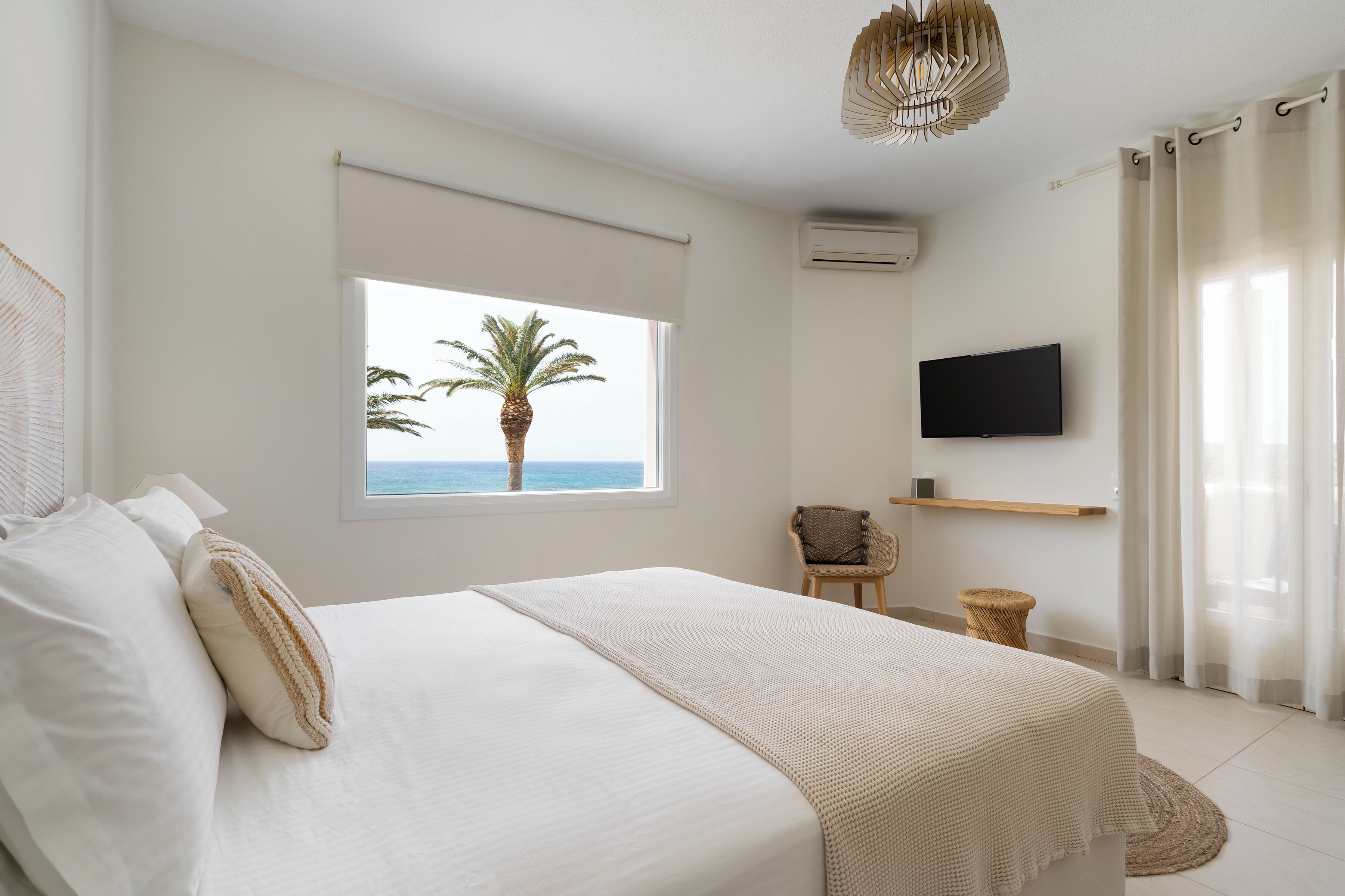 A bedroom with a bed, a window with a view of the ocean and palm tree, a chair, a television, and a ceiling light.