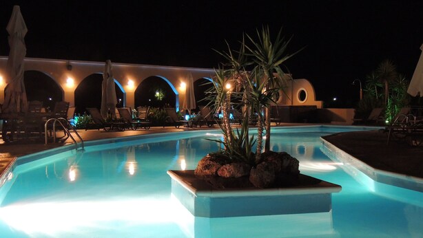 A nighttime swimming pool with a central rock feature, surrounded by lounge chairs and umbrellas, and a building with arched windows in the background. - Imerti Hotel (Lesvos)