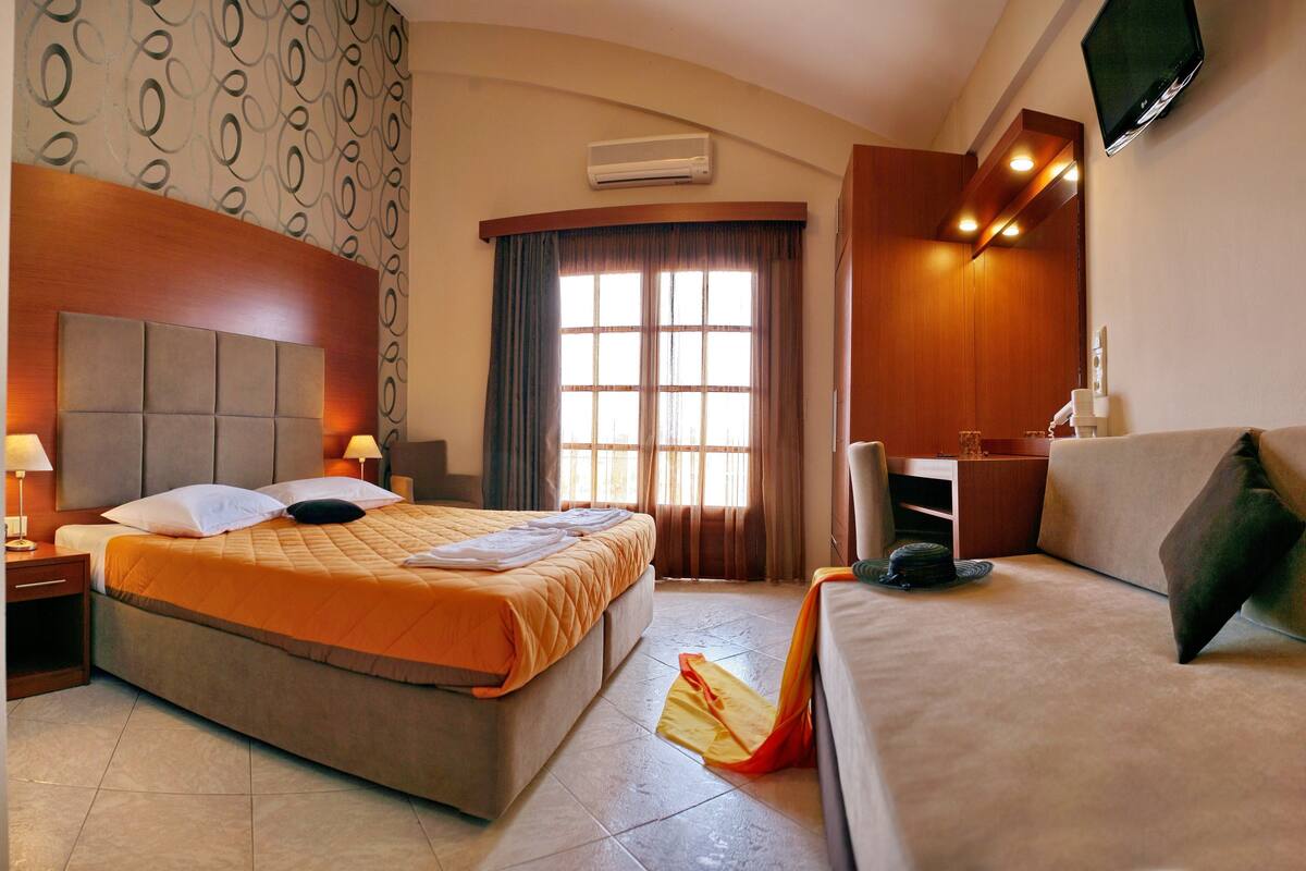 A hotel room with a bed, a sofa, a desk, a TV, and an air conditioning unit.