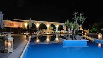 Zona de piscina con sillones de descanso y un bar, rodeada por un edificio con arcos e iluminación exterior.