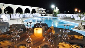 A poolside dining area with tables set for a meal, surrounded by white umbrellas and palm trees. - Imerti Hotel (Lesvos)