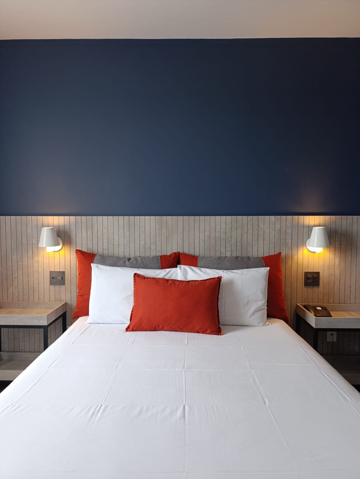 A hotel room with a bed, two bedside tables, a wooden headboard, and two wall-mounted lamps.