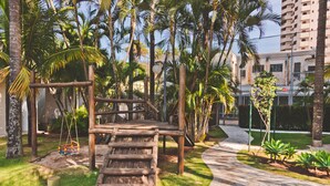 A wooden play structure in a landscaped area with palm trees and a paved walkway. - Transamerica Araraquara (Araraquara)