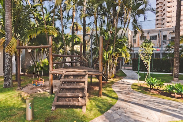 Una estructura de juego de madera en un área ajardinada, con palmeras y un sendero pavimentado.