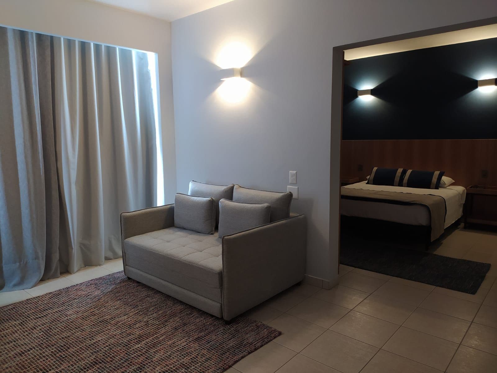 Una habitación de hotel moderna con un sofá gris, una cama con cabecera oscura y un aplique de pared.