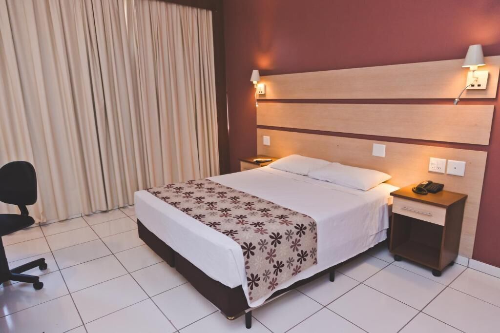 A hotel room with a bed, a nightstand, a desk lamp, a chair, and a curtain.