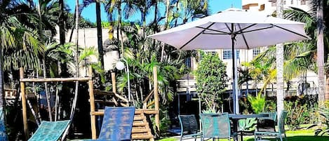 Zona de piscina con sillas de descanso y una sombrilla, rodeada de palmeras y un edificio alto al fondo.
