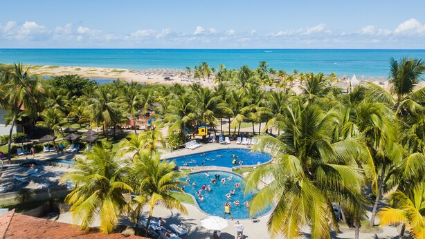 Un resort con piscina central rodeada de palmeras, una playa y vista despejada al océano.