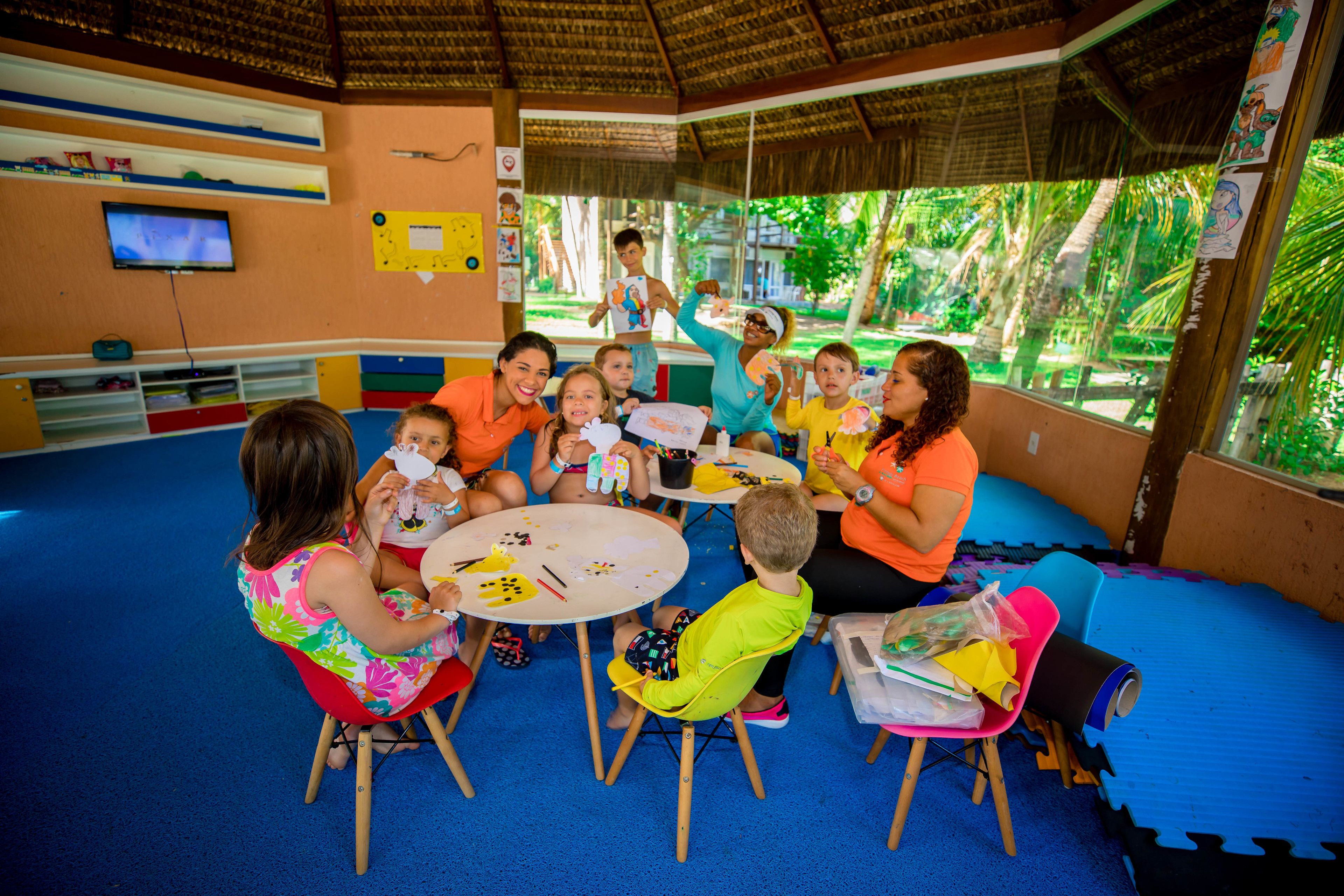 Un grupo de niños y adultos participan en una actividad alrededor de una mesa en una habitación con piso azul, muebles de madera y una ventana que ofrece vistas al exterior con vegetación.