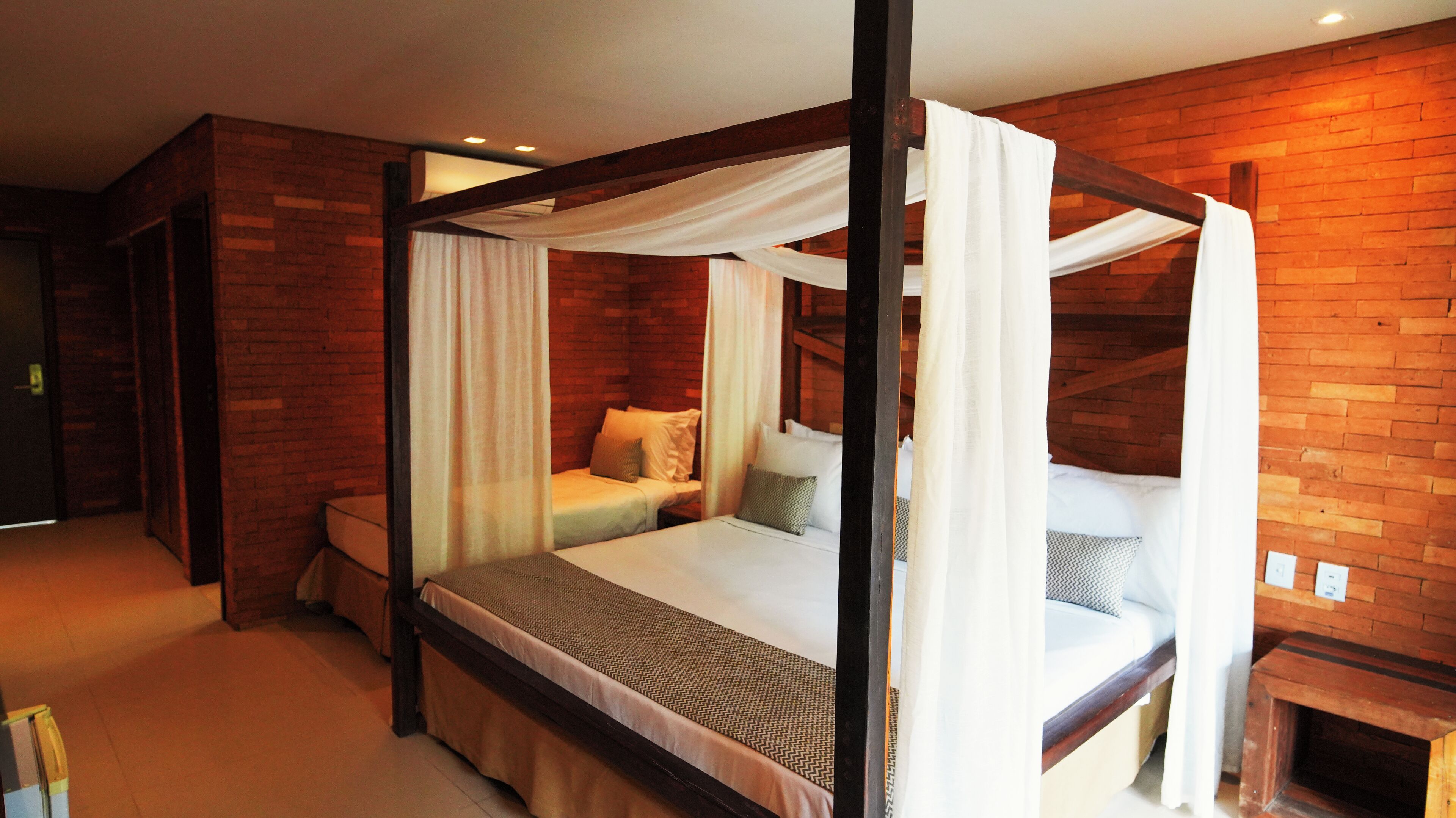 A four-poster bed with white drapes, a wooden bench, and a brick wall.