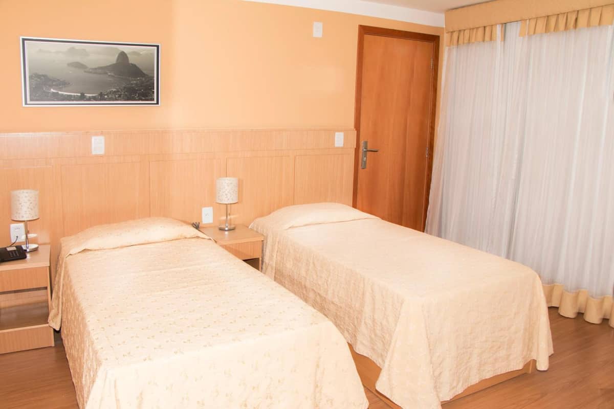 A hotel room with two beds, a wooden headboard, a nightstand with a lamp, and a door with curtains.