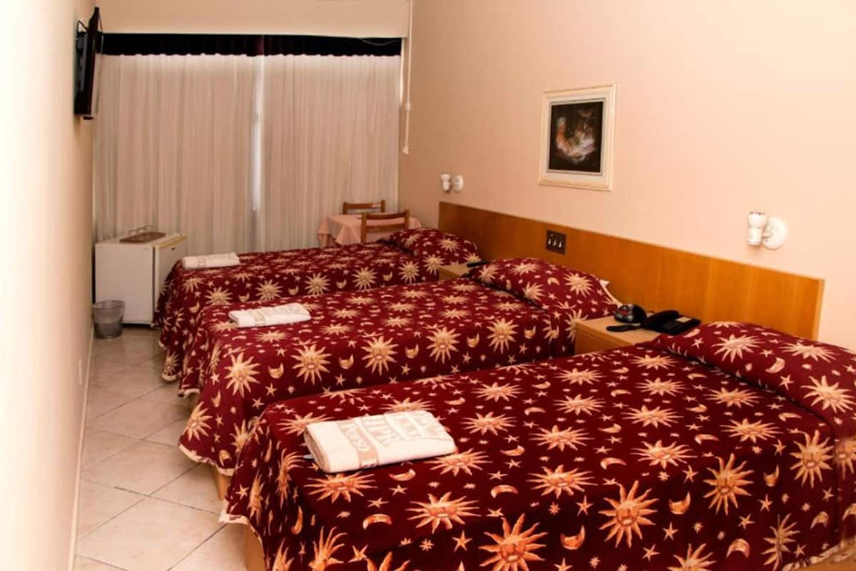 A hotel room with four beds, each with a red and gold patterned bedspread. There is a small table with a chair, a bedside table with a lamp, and a picture on the wall.